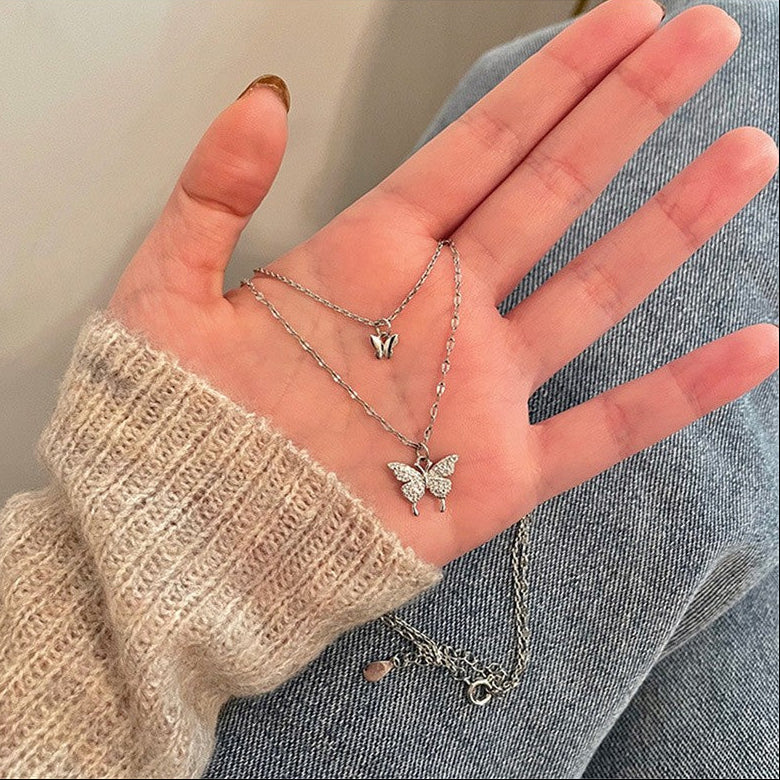Hand wearing a silver necklace with a butterfly pendant against a neutral background